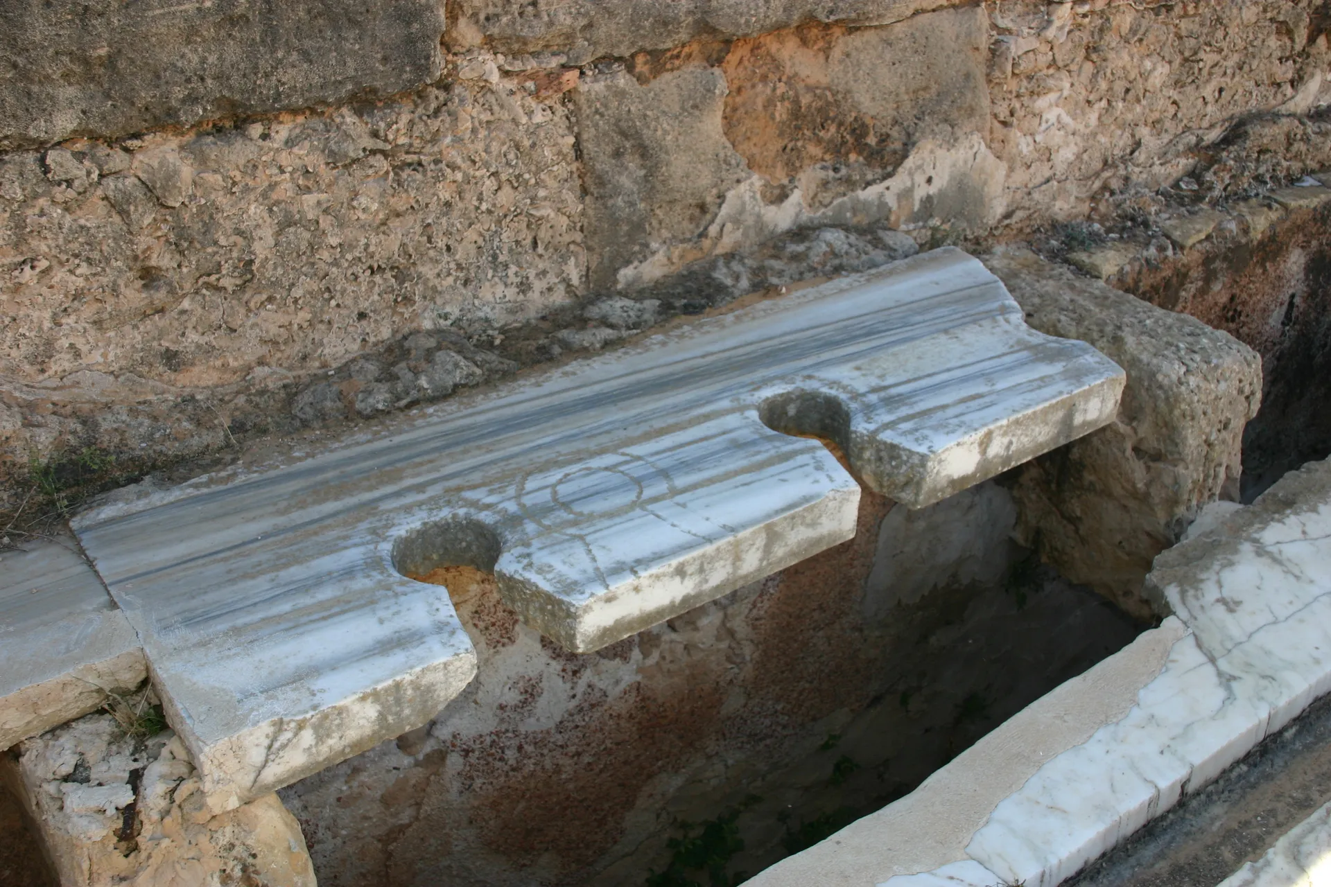 Openbare Romeinse toiletten, &eacute;&eacute;n zitplaats is foutief ingekrast [Leptis Magna]